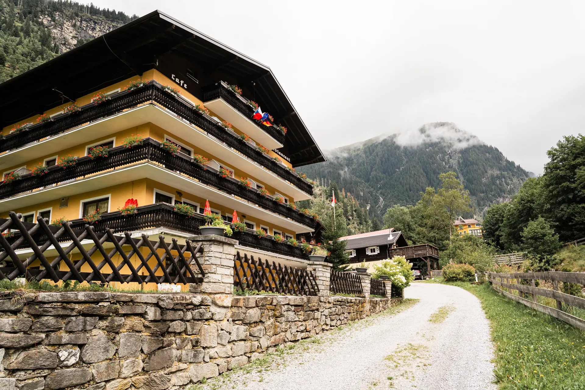Stubnerhof — rodinný hotel se žlutou fasádou a balkony plnými muškátů na kraji lesa v údolí Kötschachtal, v pozadí hora v mlze.