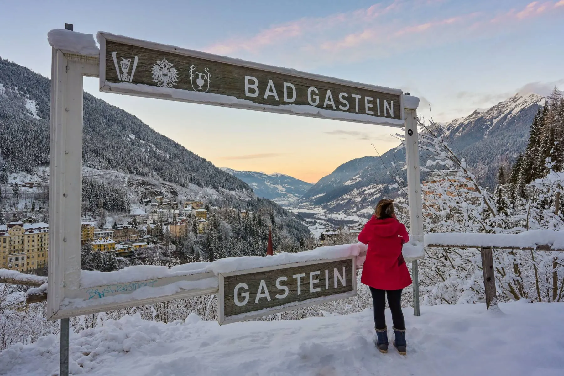 Vyřezávaný nápis BAD GASTEIN stojící v hlubokém sněhu, za ním světla města.